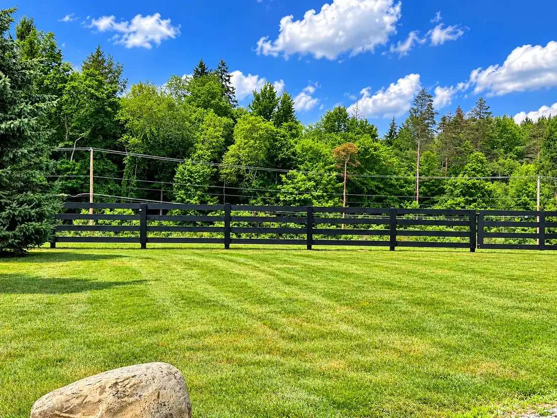Black four-board fence beside a lawn and woods